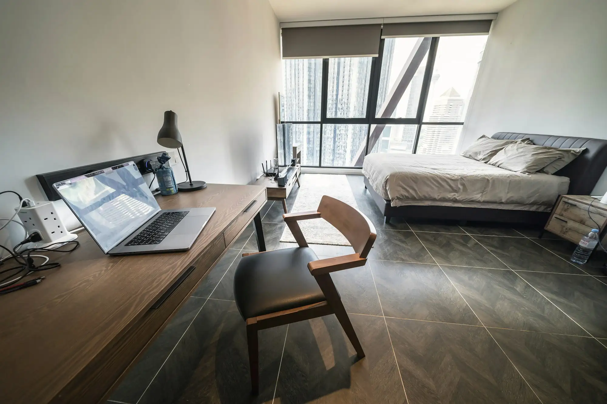 A desk with a laptop and lamp beside a bed, in a bright studio with tall windows